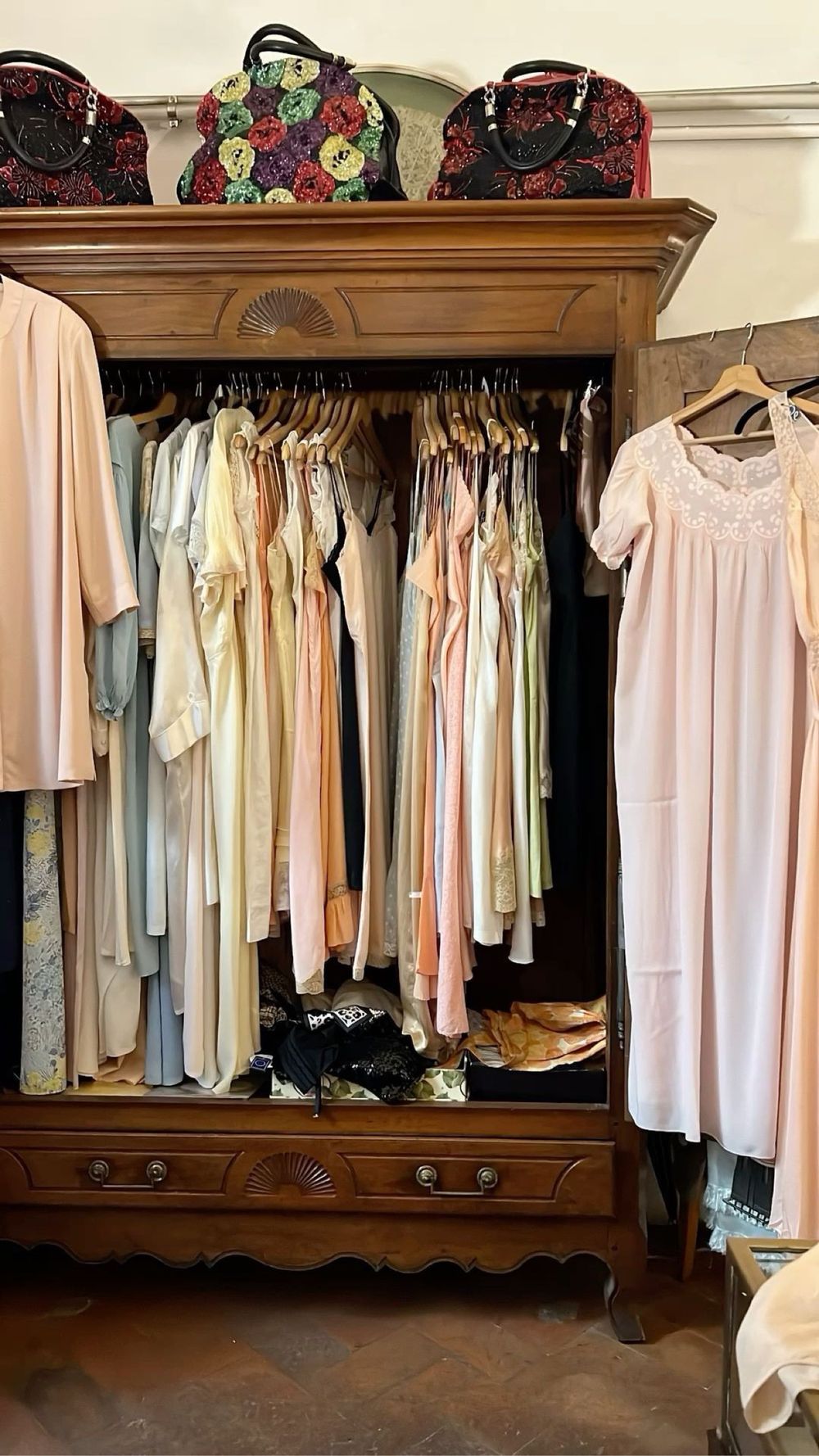 A wooden armoire filled with pastel vintage clothing, beaded bags arranged along the top